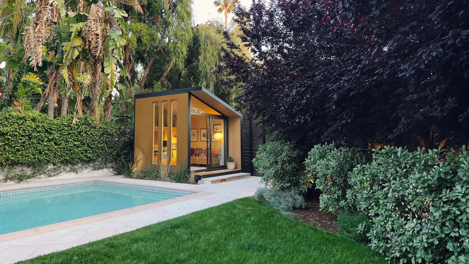 Autonomous WorkPod backyard office with ochre siding and sliding glass door next to a pool at dusk